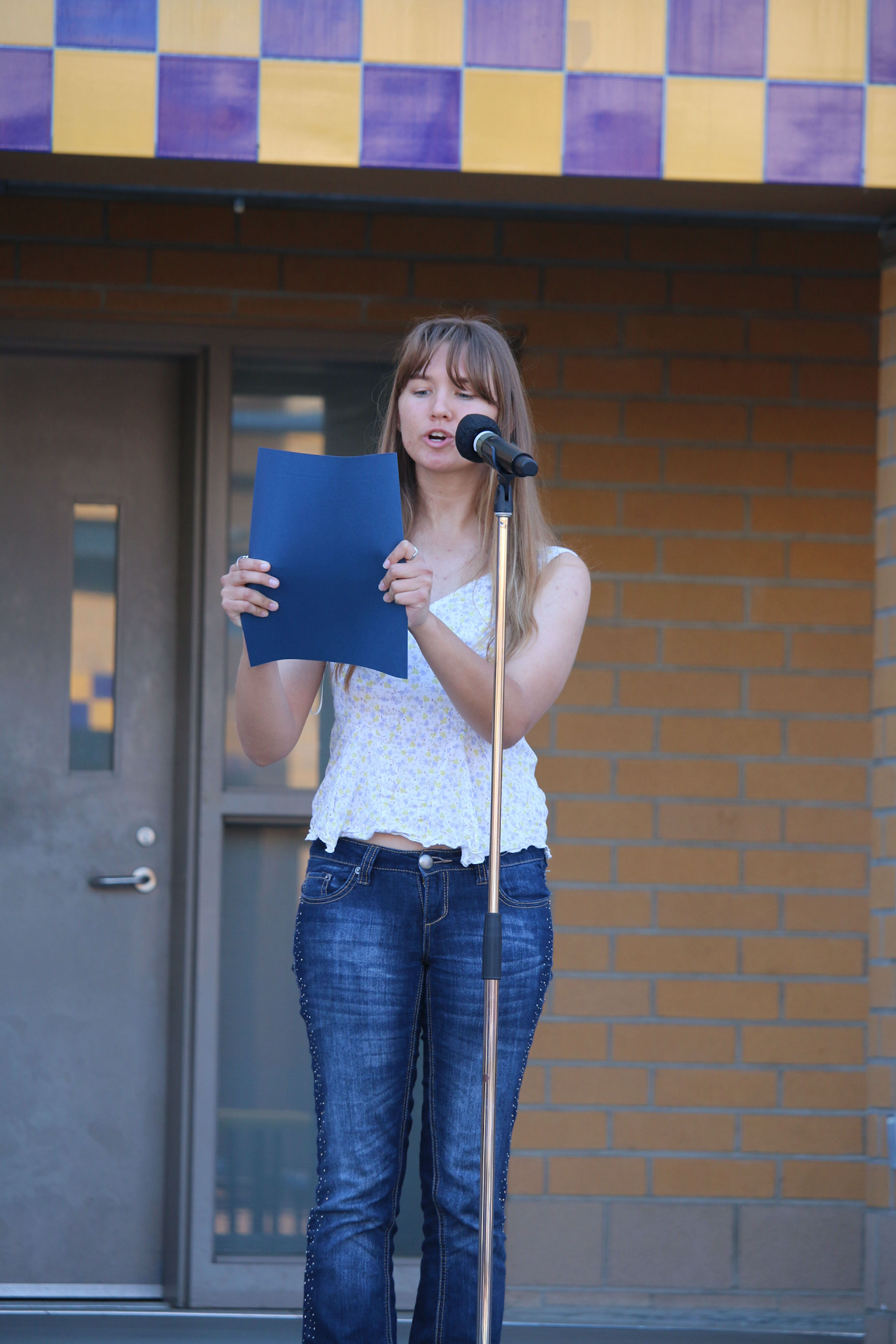 Nicole Nowak speaks into a microphone stand on the Student Center Terrace stage, holding a blue folder.