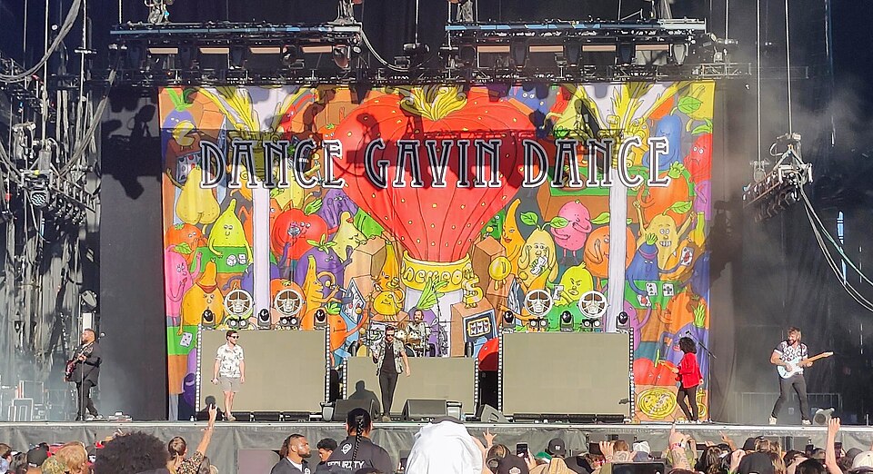 Dance Gavin Dance perform on a large outdoor stage. The stage artwork depicts anthropomorphic fruits and the words 
