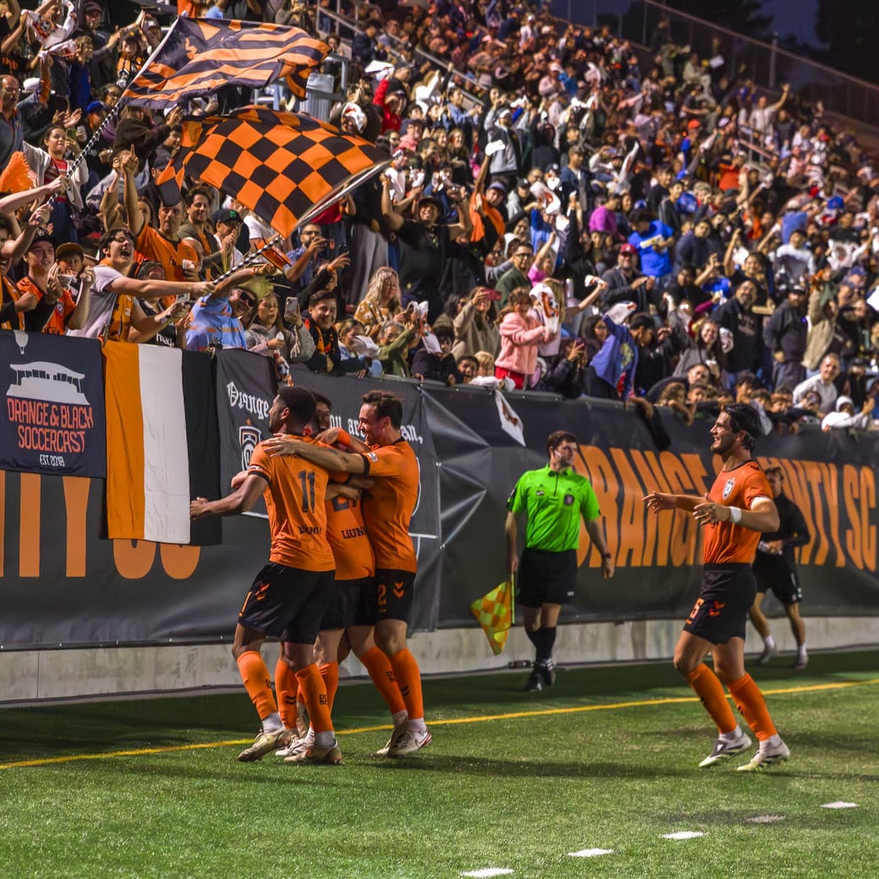 Three players in orange and black jerseys on a pitch look at fans in a seating area