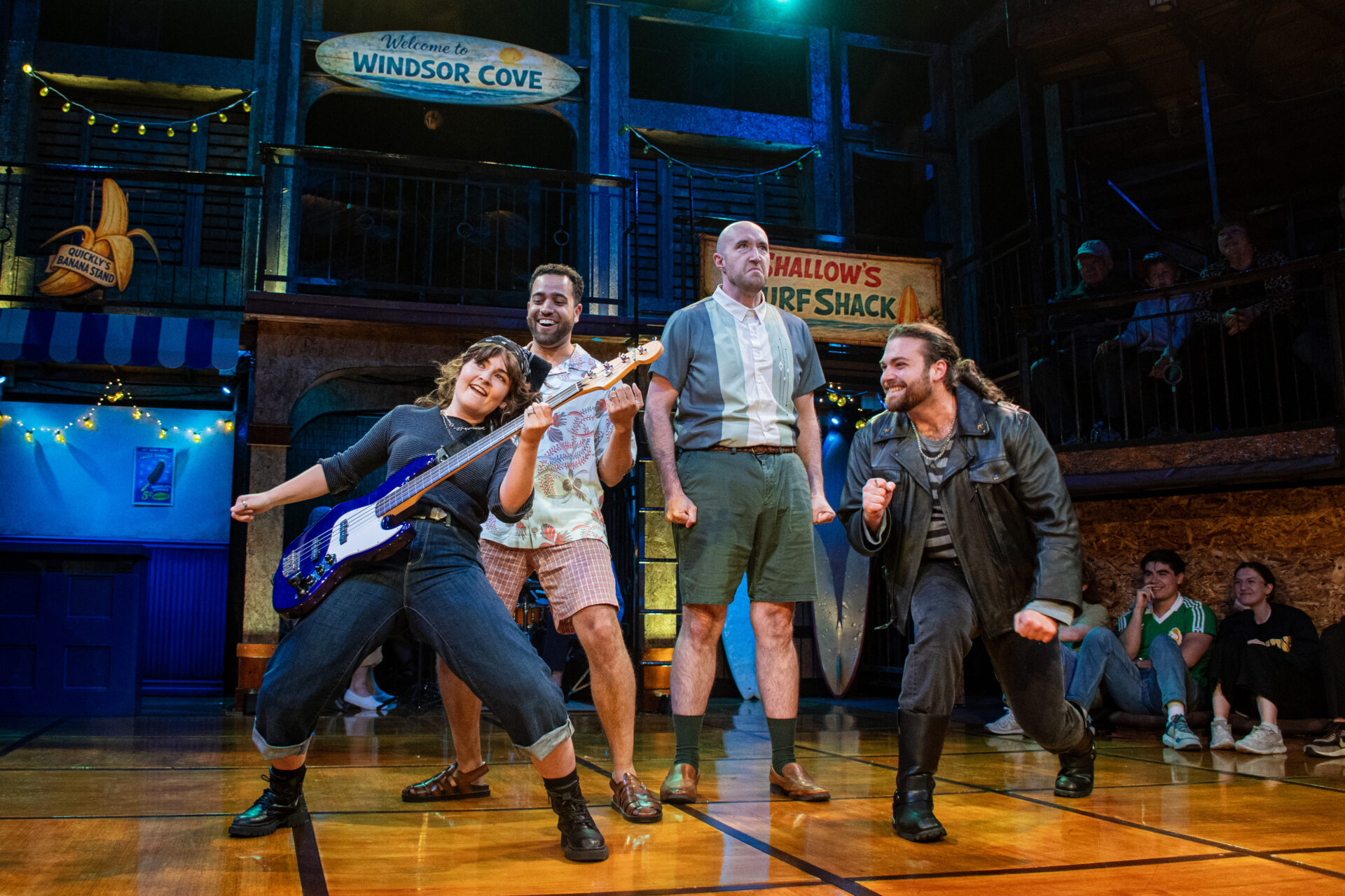 Four actors perform on a wooden stage set designed like a beachside boardwalk, with signs reading 