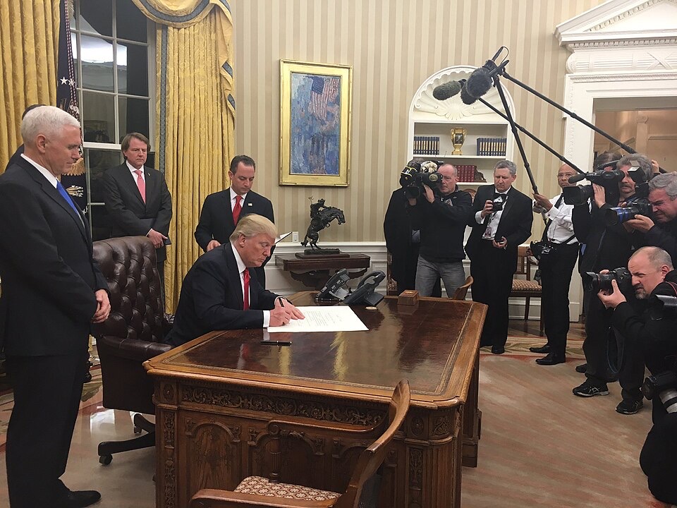 President Trump signs a paper in the Oval Office, surrounded by members of the press and White House officials
