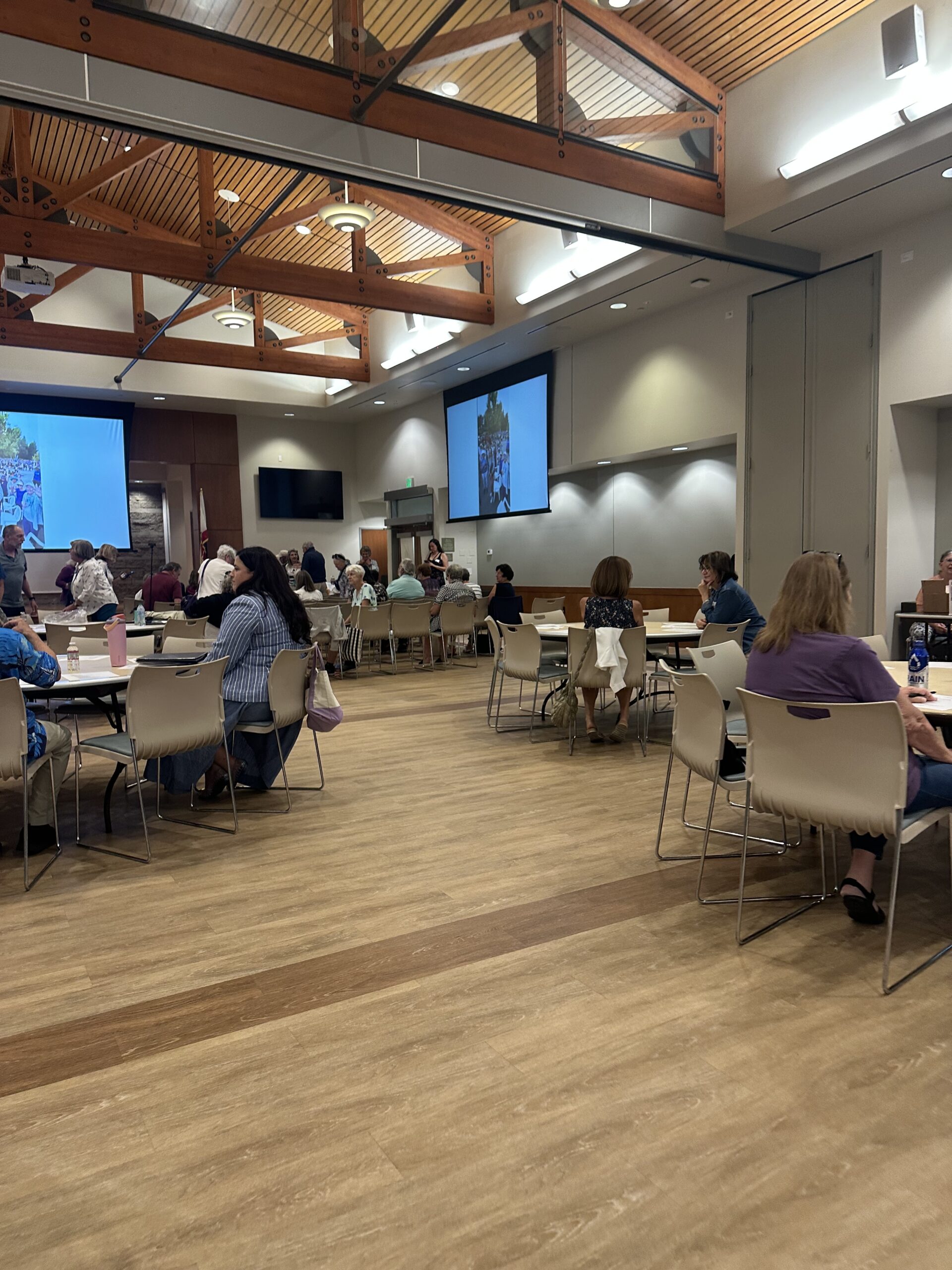 A large community meeting room with high wooden ceiling beams and skylights, where attendees sit at round tables and in rows of chairs facing two projector screens displaying photos. People are seated and standing throughout the room, some chatting in small groups.