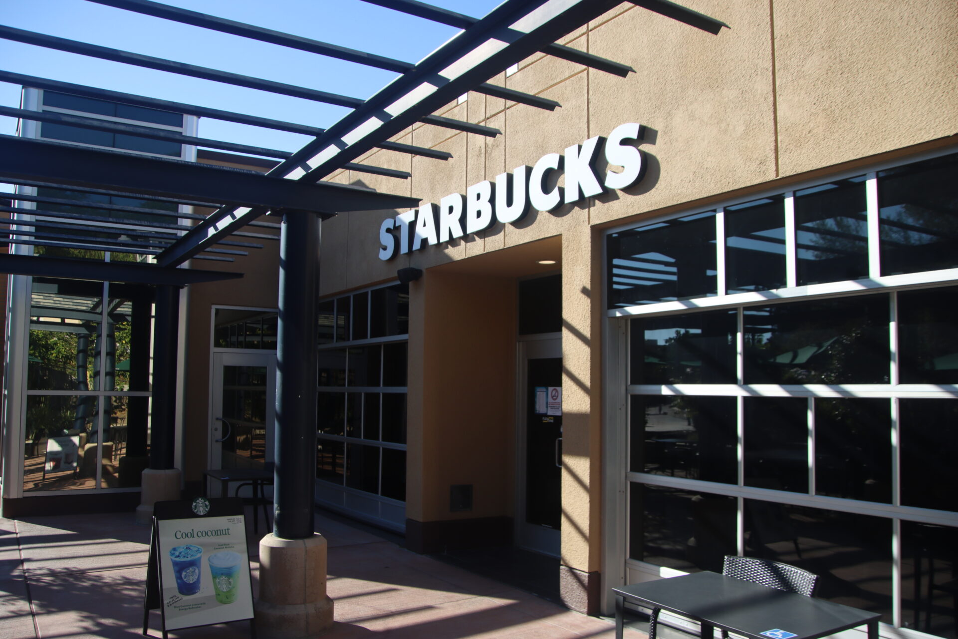 Sunny outdoor photo of a Starbucks entrance with a wooden overhead structure, an outdoor table and chairs, and a drink menu sign in front.