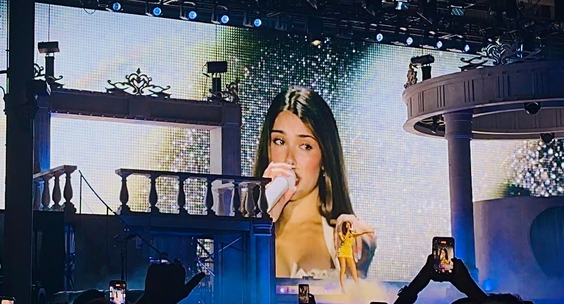 A photo of Madison Beer on a large concert screen, facing the crowd in a yellow dress while she sings into a white microphone.