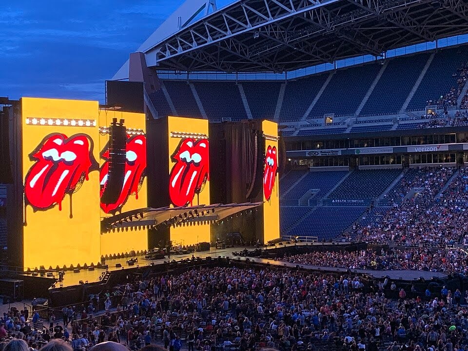A large stage and crowd with four yellow panels and four bright red mouths with tongues sticking out.