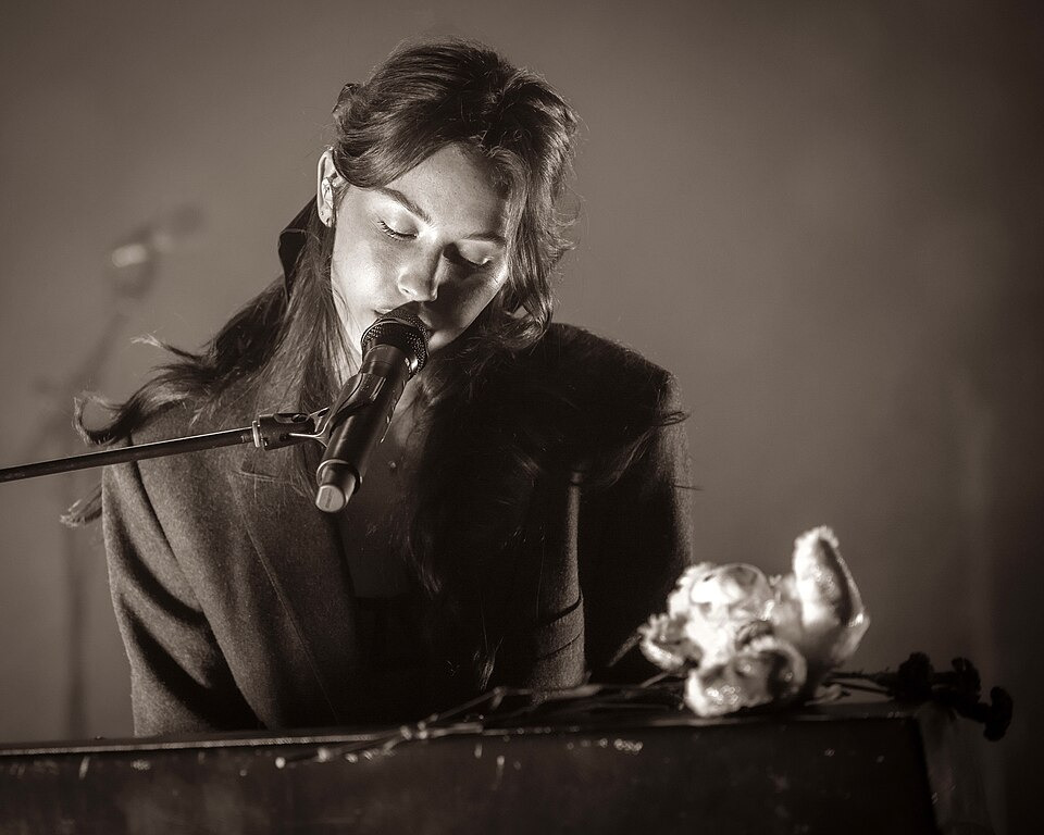 A black and white photograph of Gracie Abrams. She is sitting down, with only her upper body visible, as she plays a piano that has flowers laying on it. She is singing into a microphone as she plays with her eyes closed.