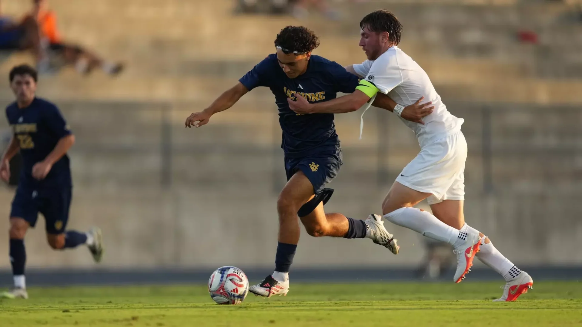 A player runs with the ball at his left foot, trying to hold off an opposing player to his left side with his left arm.