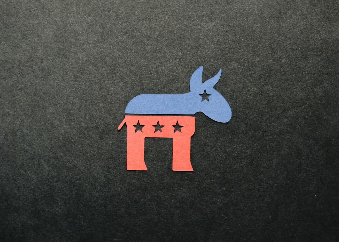 A paper cut out of the Democrat Party's donkey logo sits against a black paper background. The top half of the donkey silhouette is blue, with a single star between the donkey's eyes. The bottom half is red, with three stars lining the donkey's side.