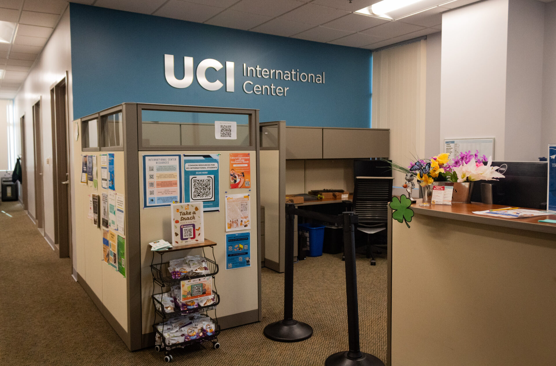 UCI International Center. A cubicle wall is shown with flyers indicating resources taped to it, and a snack rack. A desk is shown on the right with flowers on top of it.