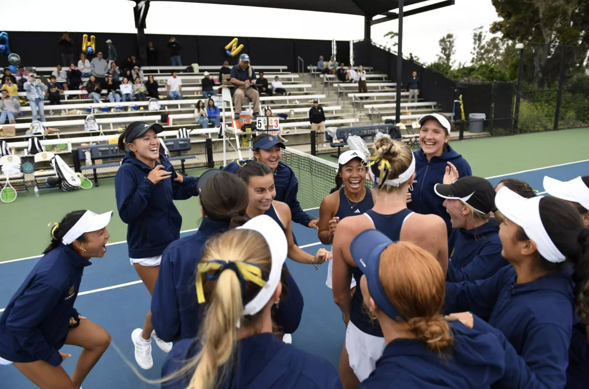 A women's tennis team is gathered at a tennis court, they're all smiling and celebrating
