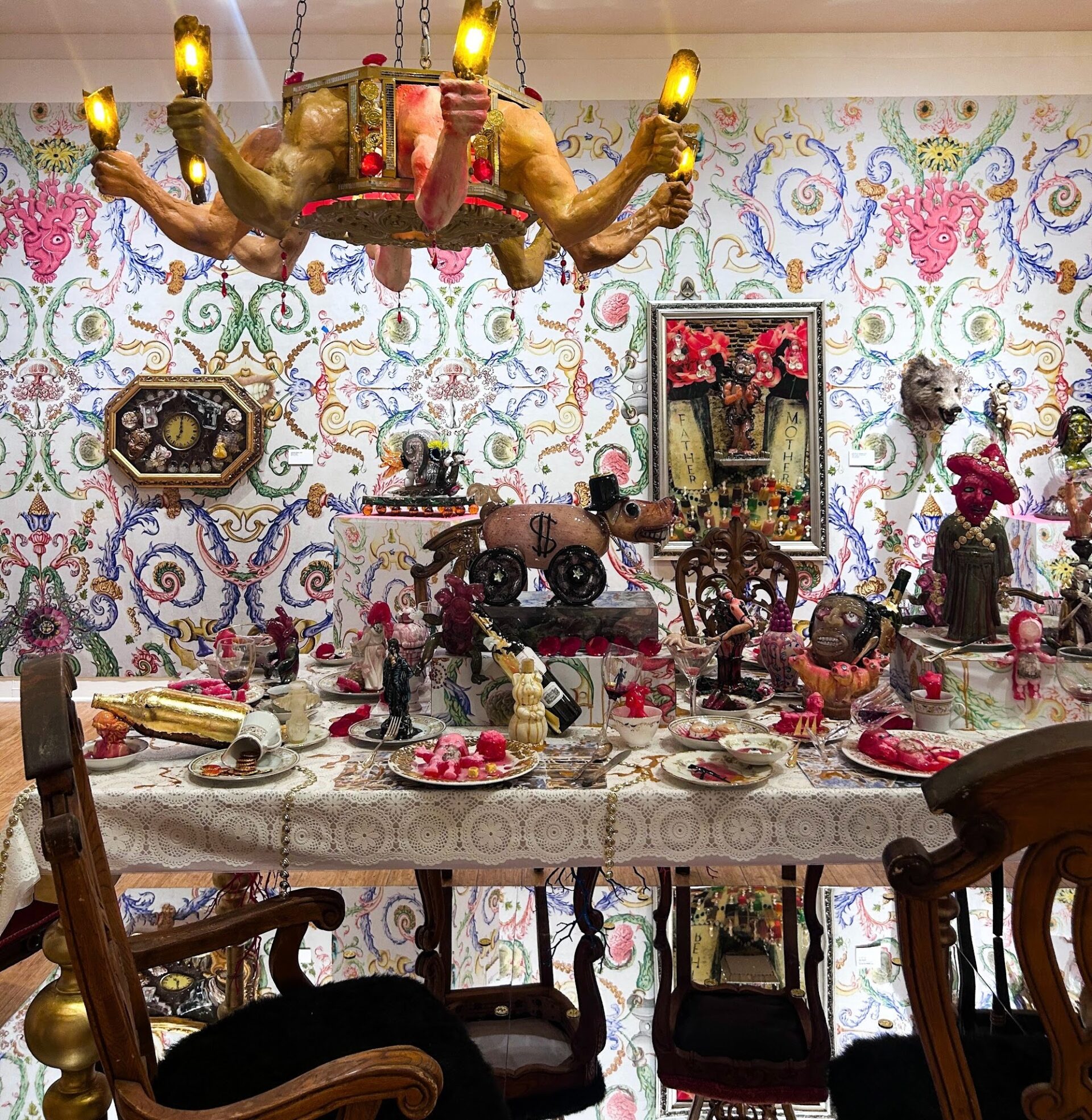 A table set with plates of food, gold coins, and a pink animal toy with a dollar sign and top hat on it. There is a large, golden chandelier and decorative wallpaper with a brain and other anatomical features as decoration.