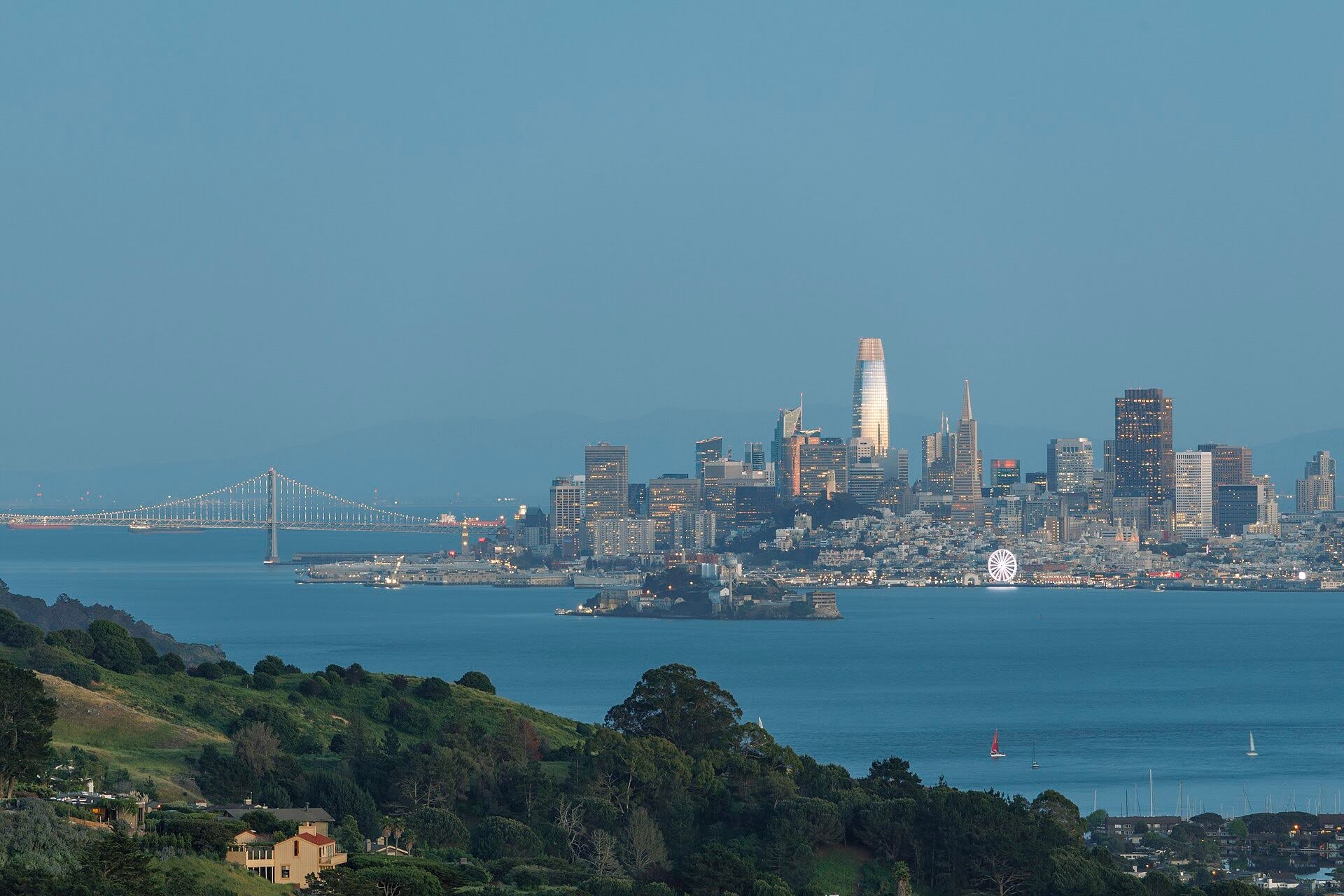 A photo of a gray bridge, the bay and San Francisco's skyline against a dusk blue sky.