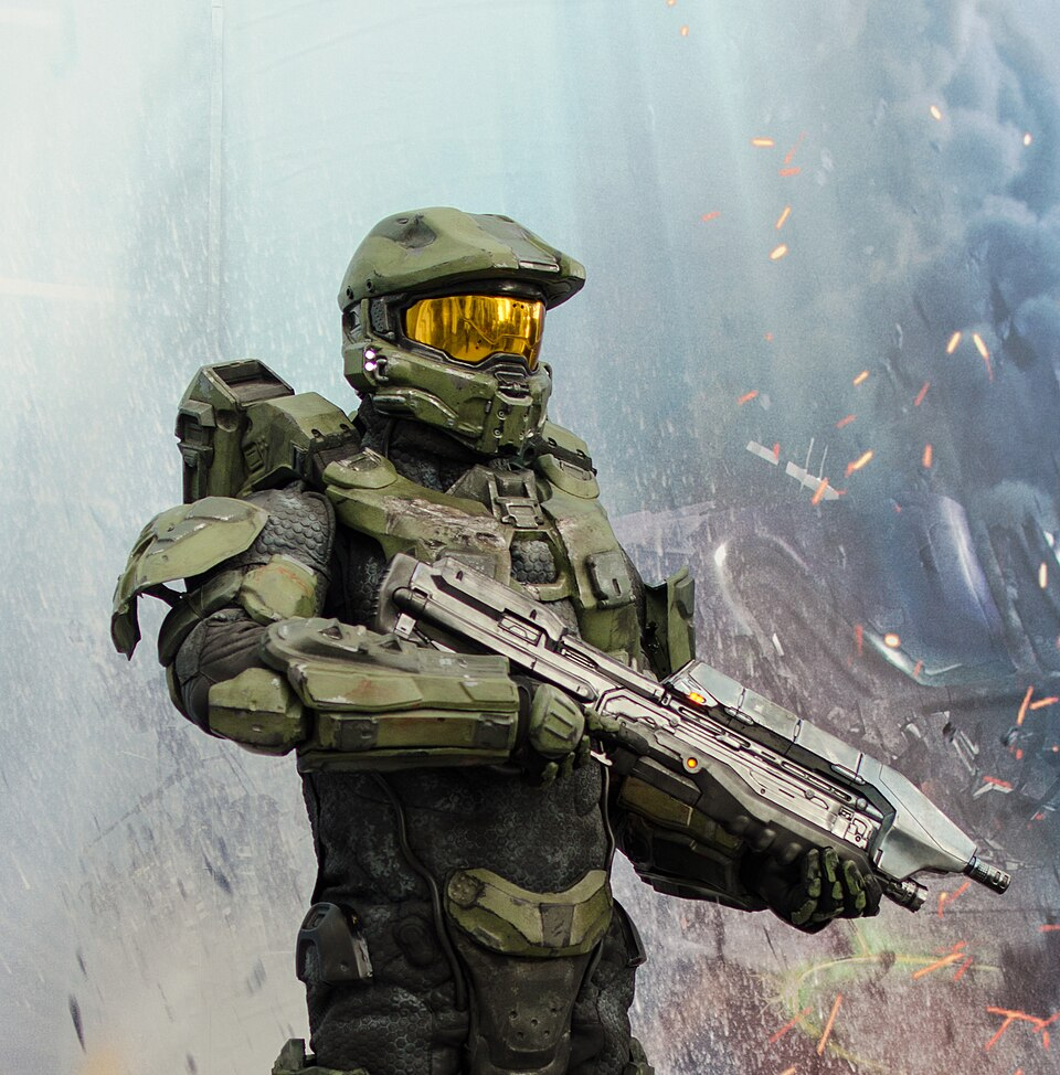 A photo of Master Chief from the Halo franchise. He is wearing camo-colored military armor and helmet, which has a yellow visor. He is holding a gun, with the wreckage of a vehicle behind him.