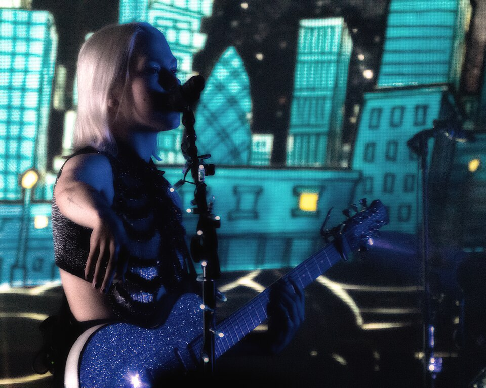 Phoebe Bridgers stands to the left of the image, playing the guitar as she sings into the microphone in front of her. Behind her, there is a nighttime, star-speckled cityscape painted cyan blue.