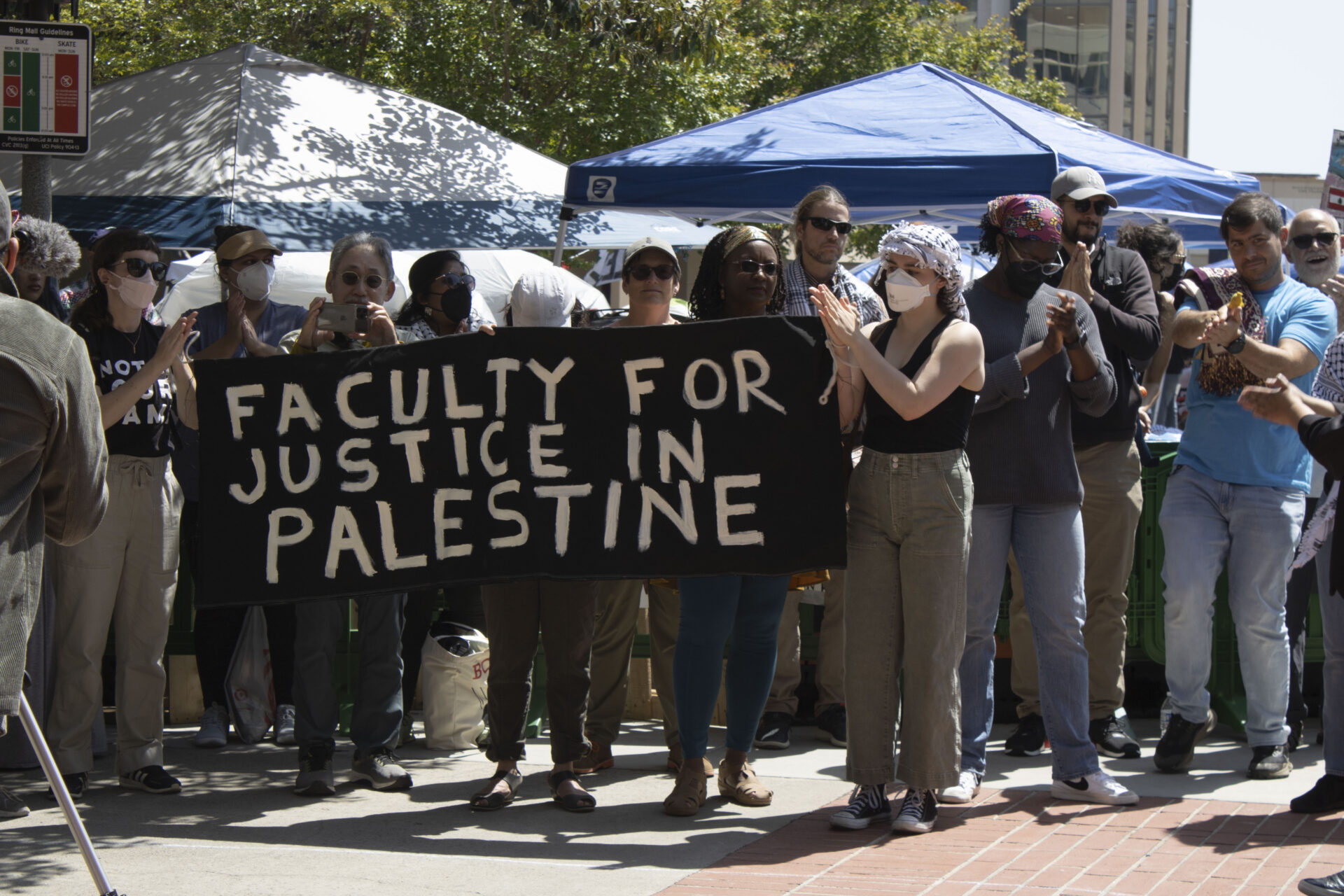 UCI faculty members stand in front of the encampment's tents, holding a black banner that reads 