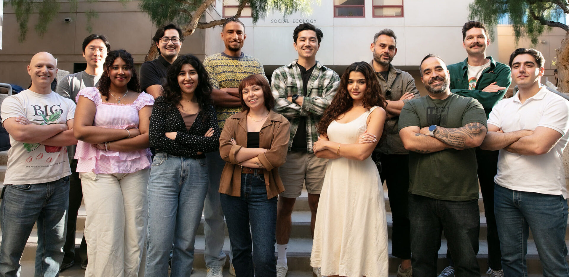 A group photo of thirteen people standing on outdoor steps in front of a building labeled 