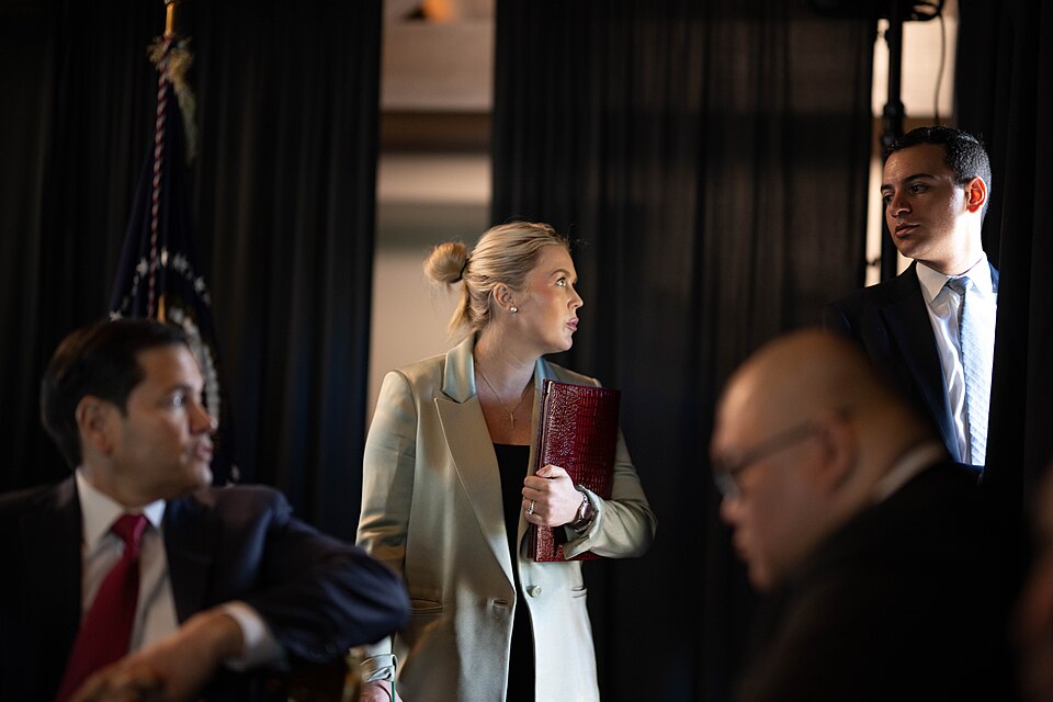 Karoline Leavitt stands in the middle of the frame with her hair in a bun and a red snakeskin folder tucked under her arm. She is looking to her left and speaking to a man at her side, while Marco Rubio and another man sit in the foreground.
