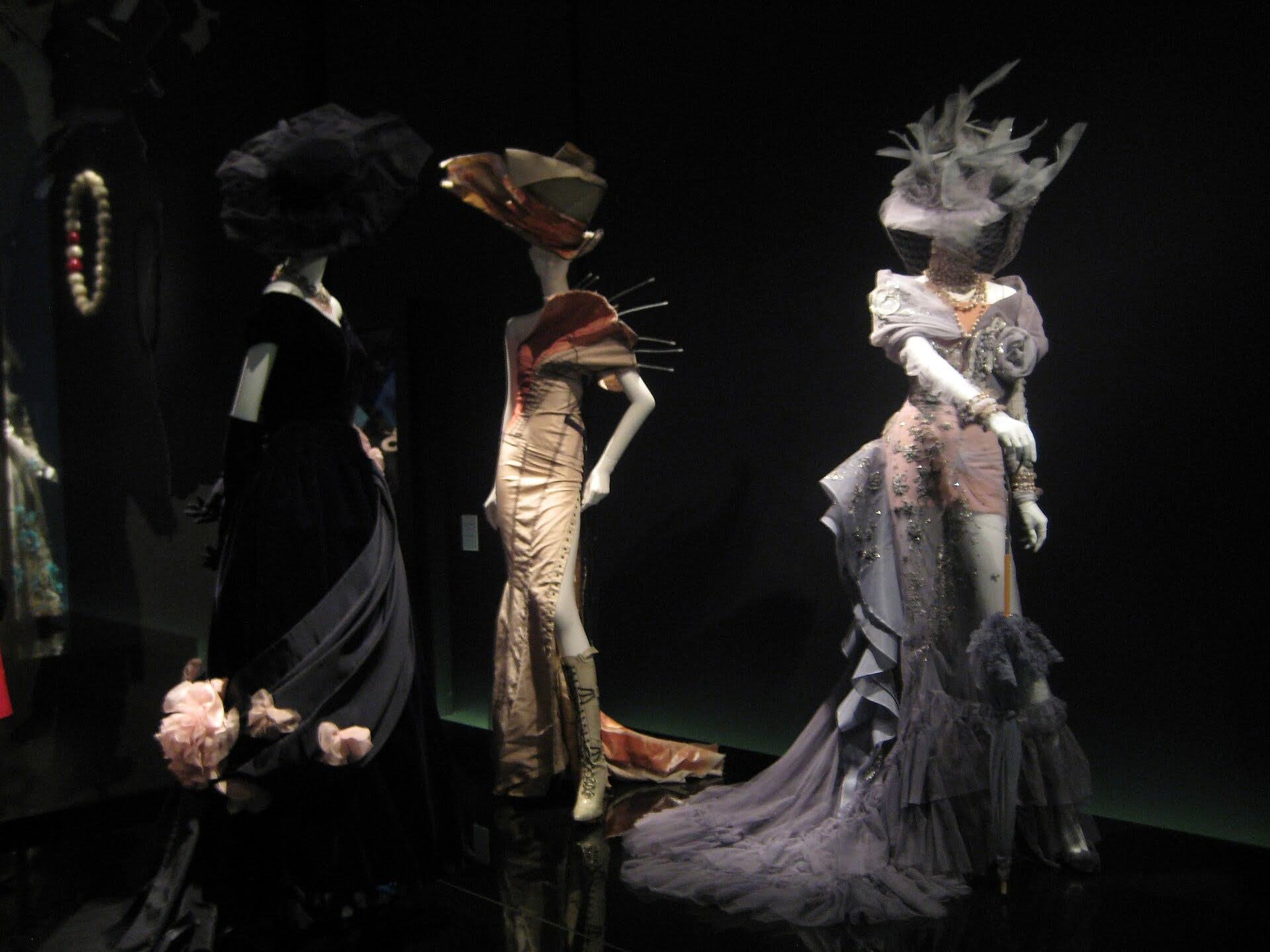 Three white mannequins against a black background, one wears a sleek black dress, another wears a pinned creme dress, and the third wears a ruffly silver dress.