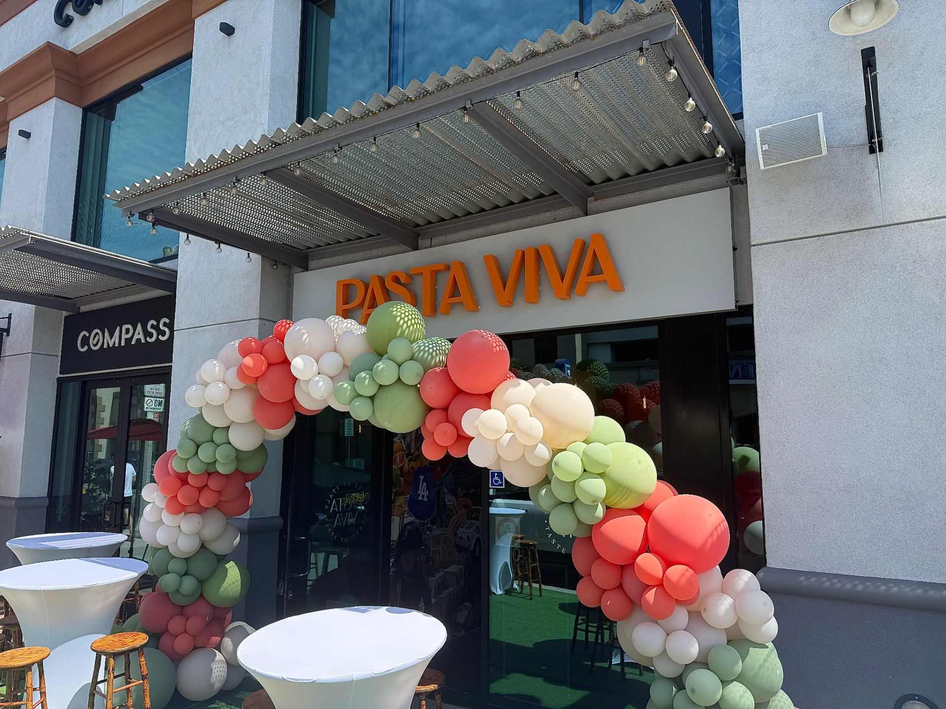 A photograph of Pasta Viva's storefront, where the restaurant's name is written along the top of the building in orange, all capital lettering. In front of the restaurant, there is a single balloon arch colored green, red and white, as well as seating scattered around.