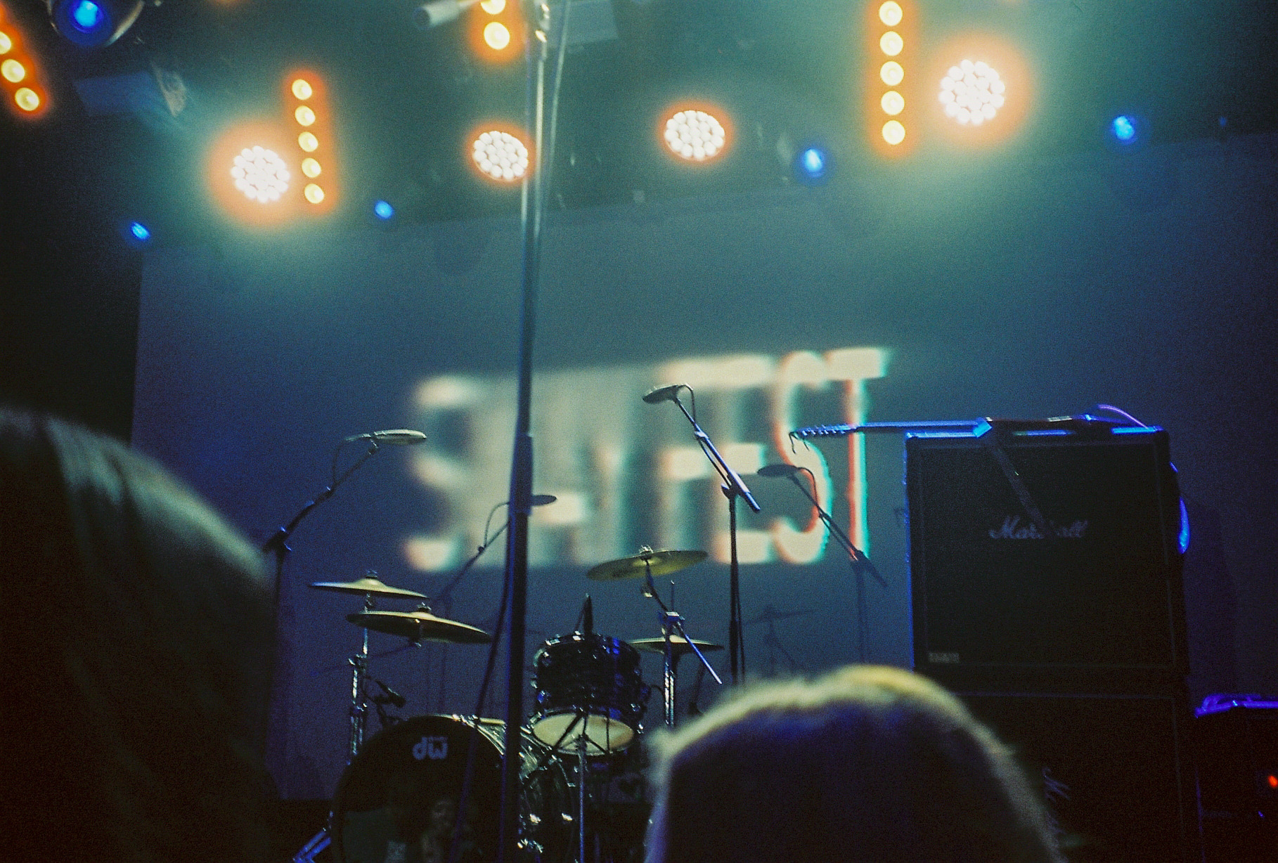 A concert stage with a set of drums, speakers and microphones. The stage is being lit with overhead stage lights, with the words 