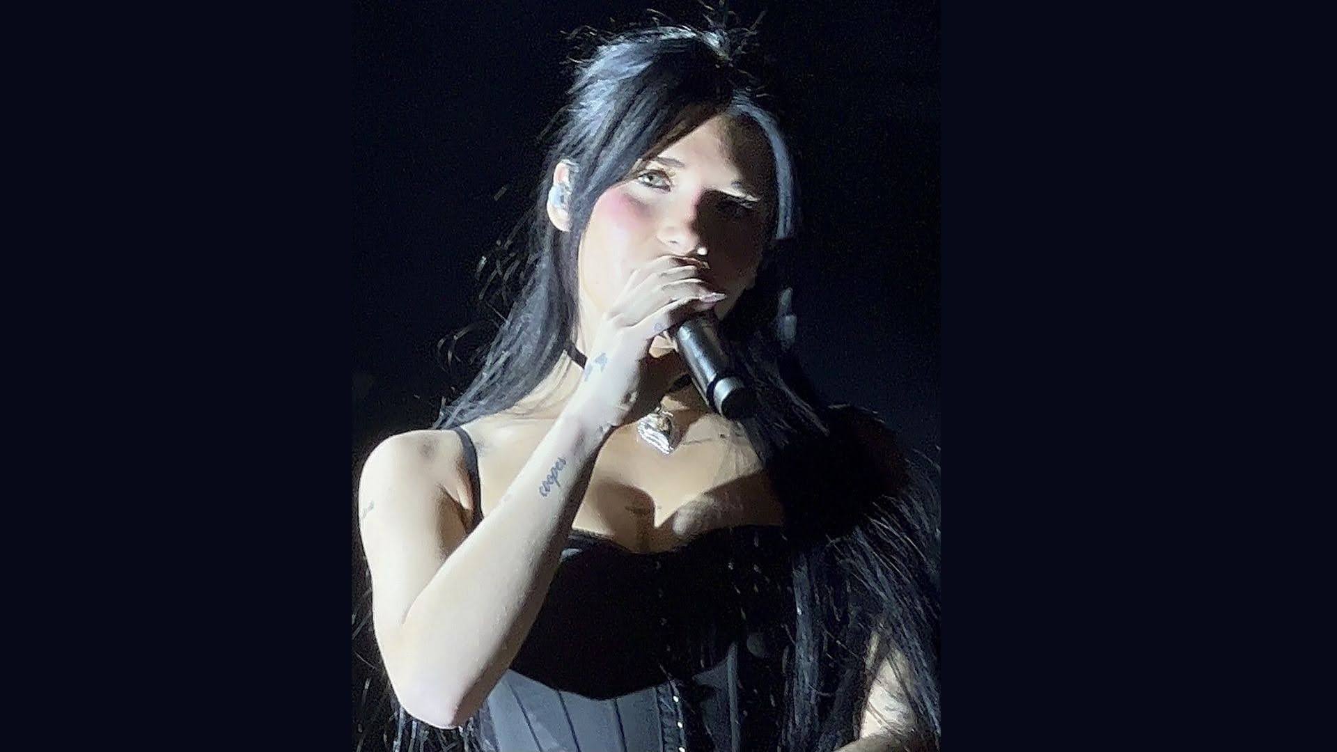 A woman holds a black mic to her mouth against a blank, black background. She has dark black hair and is wearing a black corset-styled top; her head is tilted upward to the left as she looks forward.