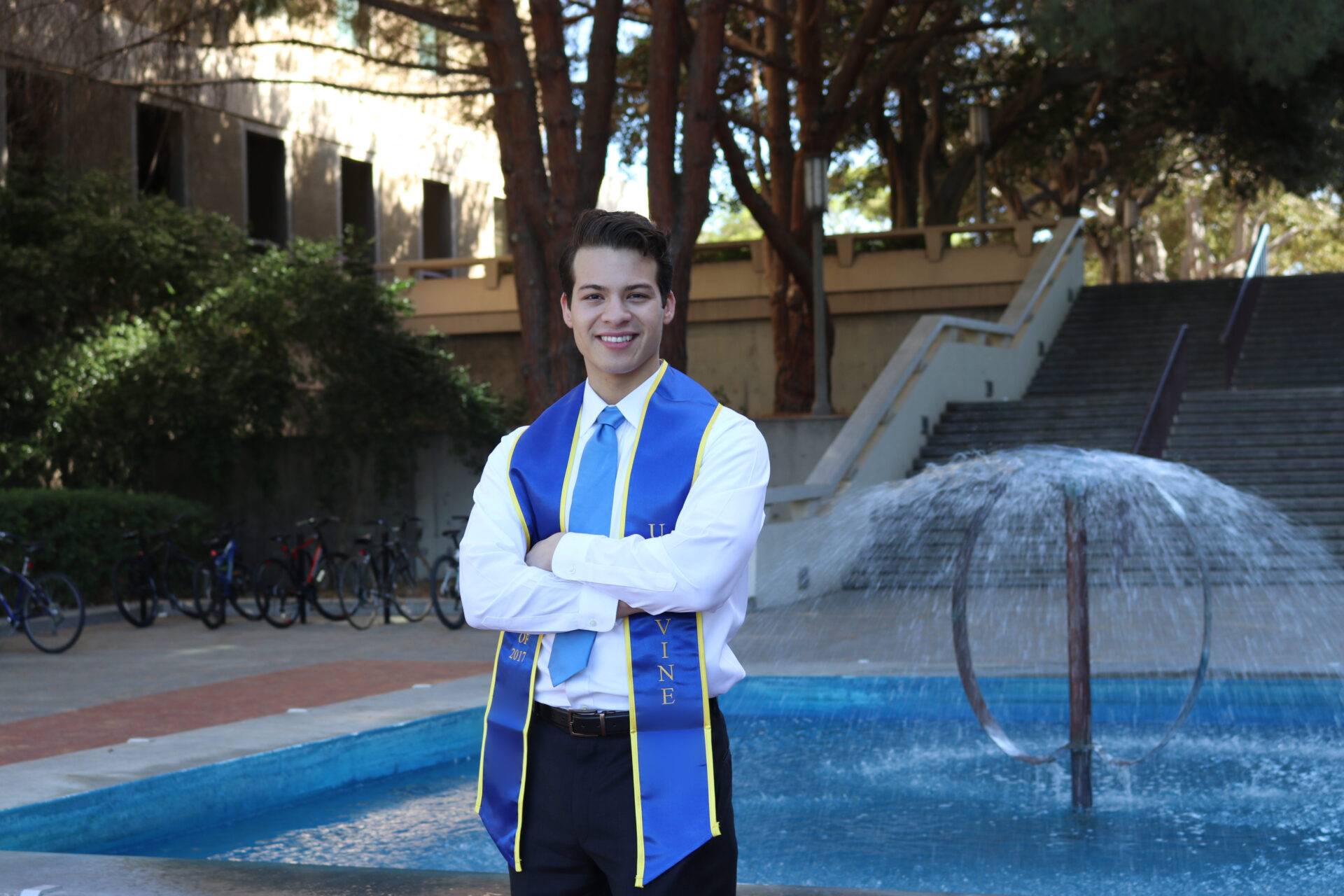 A smiling graduate wearing a blue and gold UC Irvine 2017 stole stands in front of a campus fountain.