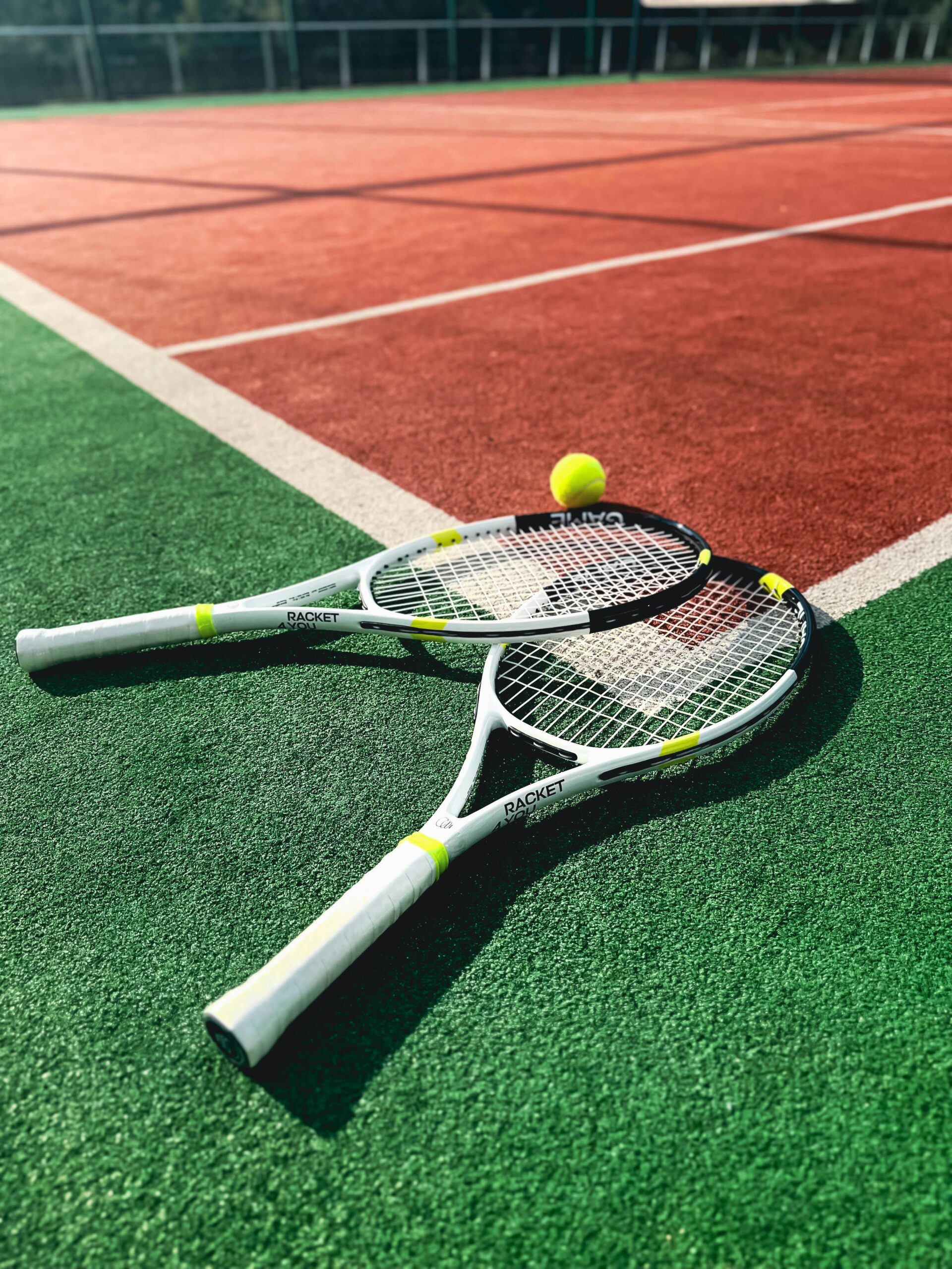 Two tennis raquets, one places slightly on top of the other, on the floor of the tennis courts with a tennis ball right beside them
