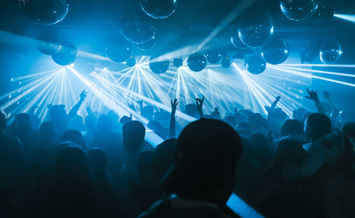 A crowd parties in a room lit up by strobe lights and filled with balloons. The crowd is cheering and dancing.